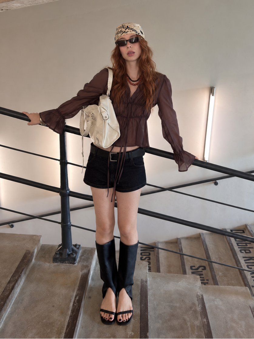 The Cocoa Smoke Ethereal Drawstring Sheer Blouse styled with black denim shorts on a contemporary staircase.