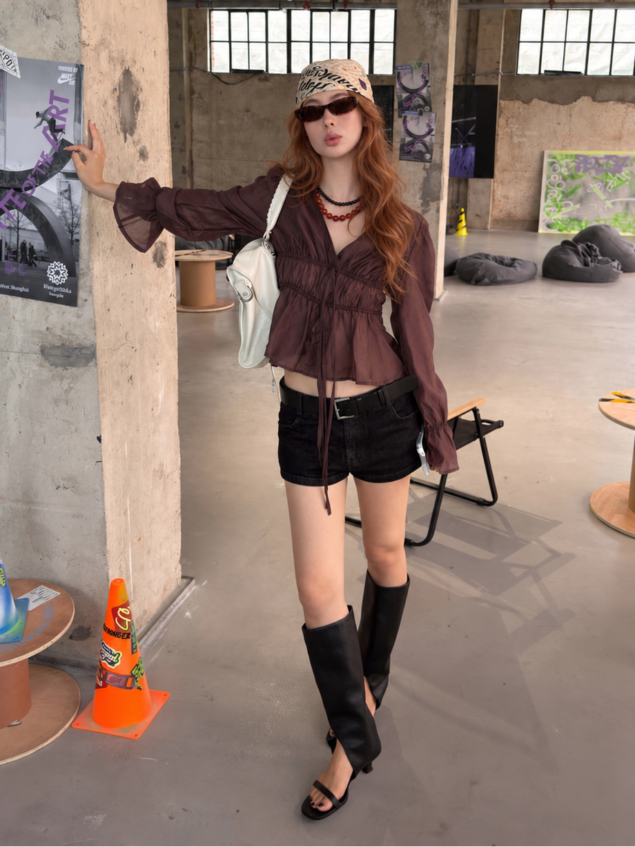 Full-body styling of the Cocoa Smoke Ethereal Drawstring Sheer Blouse paired with denim shorts and knee-high boots.
