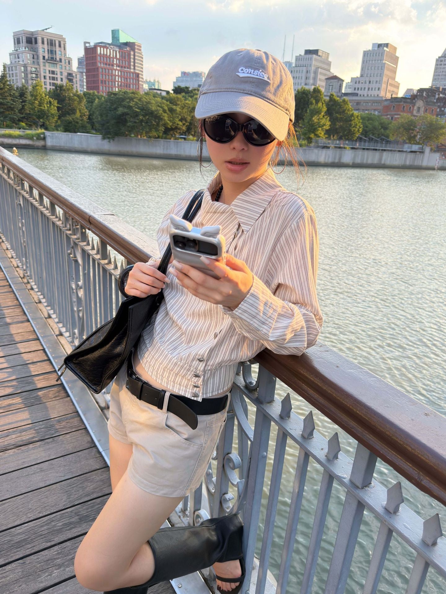Woman wearing an asymmetric collar striped cropped cotton shirt while checking her phone on a riverside bridge