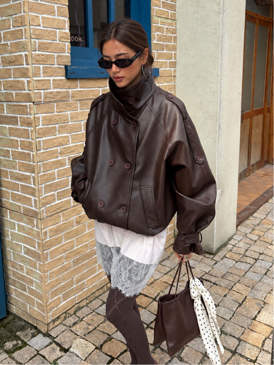 Woman wearing the Avery Oversized Faux Leather Moto Jacket in Espresso Brown with the stand collar fastened, styled with a lace dress and sheer tights.