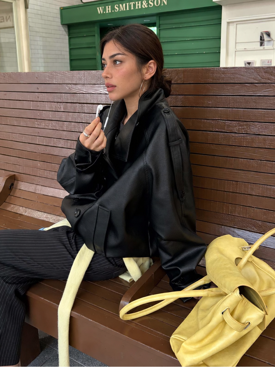 Side view of the Avery Oversized Faux Leather Moto Jacket in Midnight Black styled with pinstripe trousers while seated on a wooden bench.