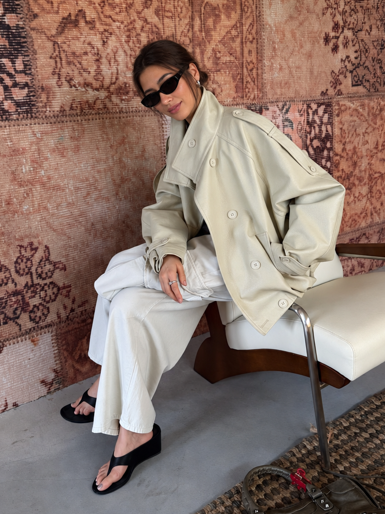 Woman wearing the Avery Oversized Faux Leather Moto Jacket in Soft Ivory while seated in a lounge chair, styled with wide-leg white trousers.